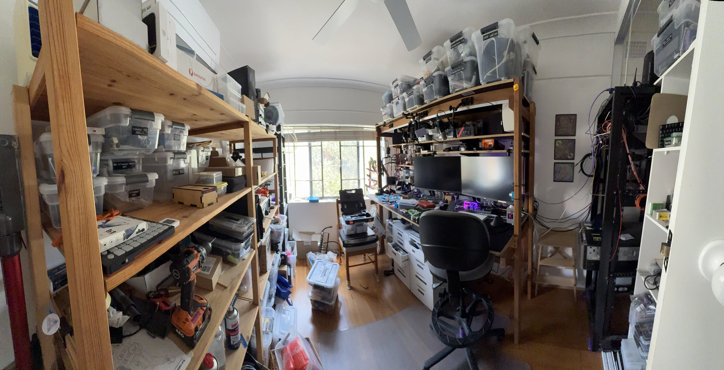 The project room and my workbench A panorama of a small room with shelves on the left, window centre, and a workbench on the right, with server rack further right. There's many transparent boxes and misc equipment sitting on the shelves.